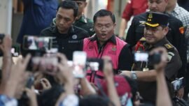 Mantan pejabat yang mengenakan rompi tahanan berwarna merah muda dikawal aparat penegak hukum di tengah kerumunan awak media dan warga yang merekam menggunakan telepon genggam.